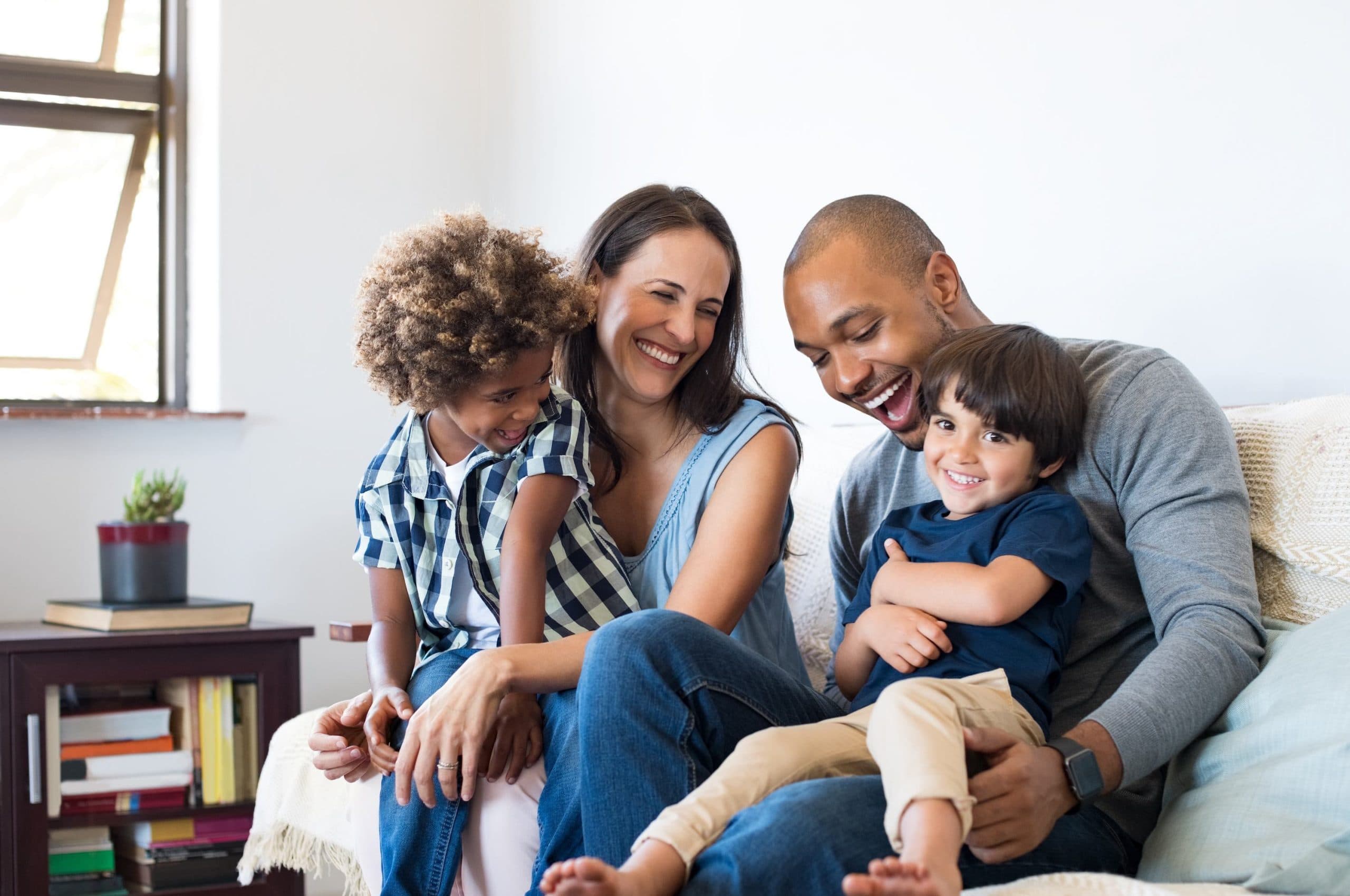 Two parents and their two young children laughing together on the couch in the living room of their home