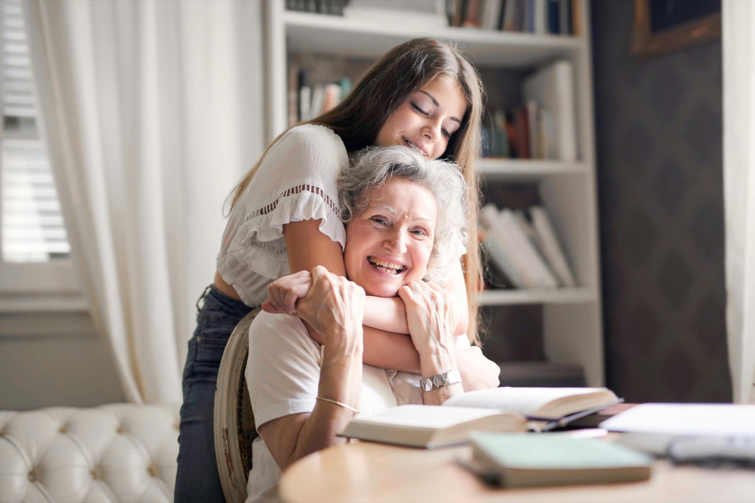 A grandmother laughing at a desk of open books while her granddaughter hugs her from behind