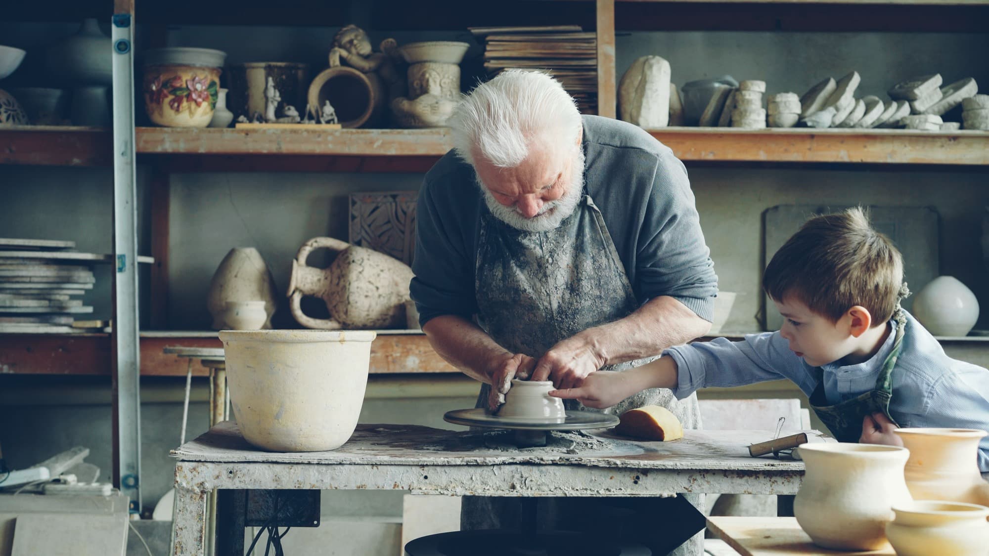 An older potter guides a boy's hands at the wheel in a workshop lined with pots