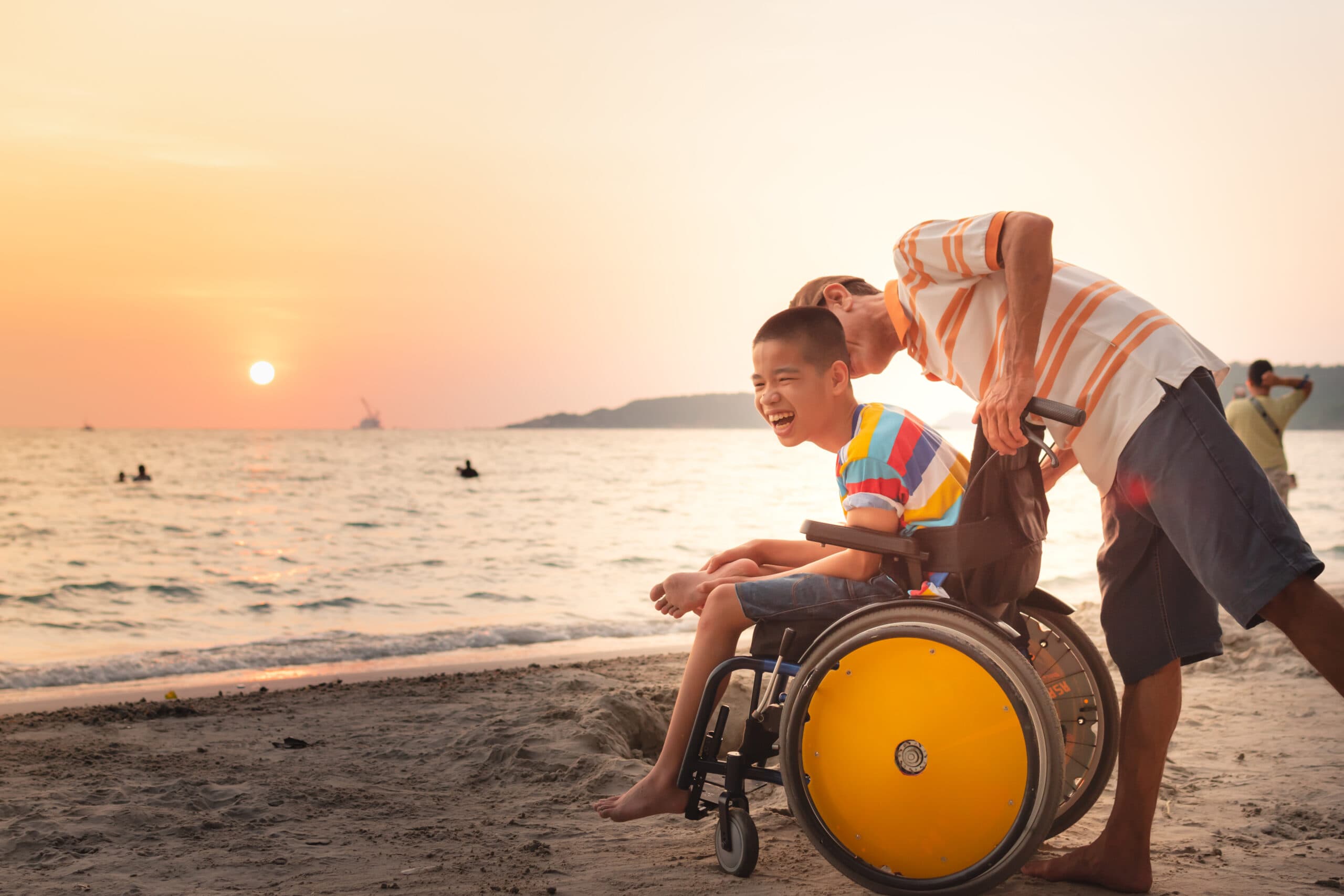 A boy in a wheelchair at the water's edge at sunset, laughing, with an adult crouched beside him